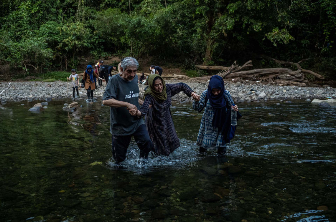 A group of older Afghan adults, exhausted after several days of crossing the jungle, help each other cross a river. © Federico Rios Escobar